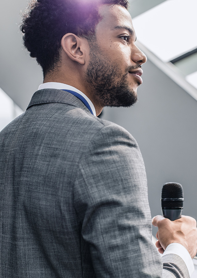Man wearing a grey suit and holding a microphone facing the right hand side of the image. 